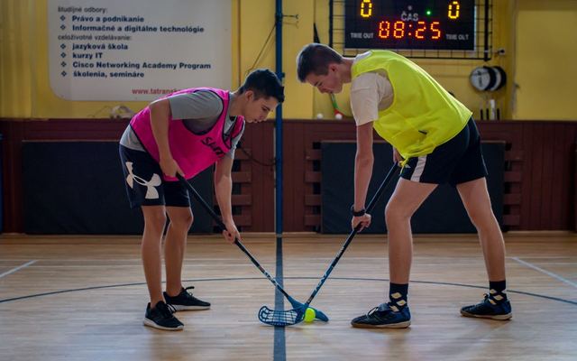 Two teenagers face off in a floorball match inside a sports gym with equipment visible in the background.