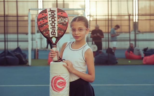 A young girl with glasses holds a paddle racket on an outdoor tennis court during the day.