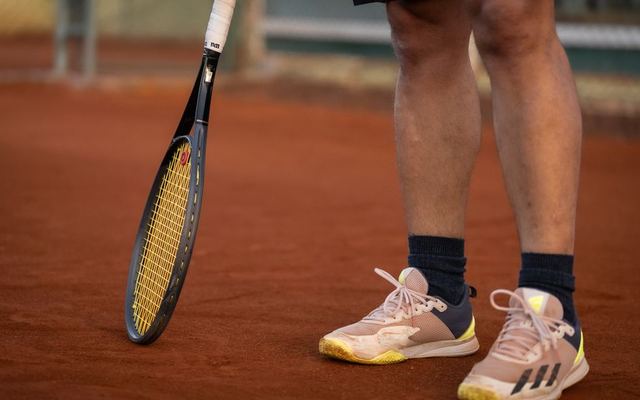 A close-up shot featuring a tennis racket and player's shoes on a clay tennis court.